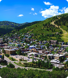 Mental Health Advocacy Park City A small town with clustered buildings nestled in a green, hilly landscape, surrounded by trees under a bright blue sky with a few white clouds.