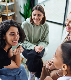 Mental Health Advocates Park City Four women sit in a circle indoors, laughing and smiling while having a conversation. The setting appears casual and friendly, with natural light and plants in the background.