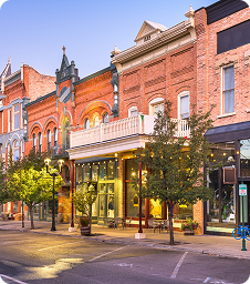 Park City Community Partnerships Historic brick buildings with large windows and decorative facades line a quiet downtown street at dusk; trees, storefronts, and a bike rack are visible along the sidewalk.