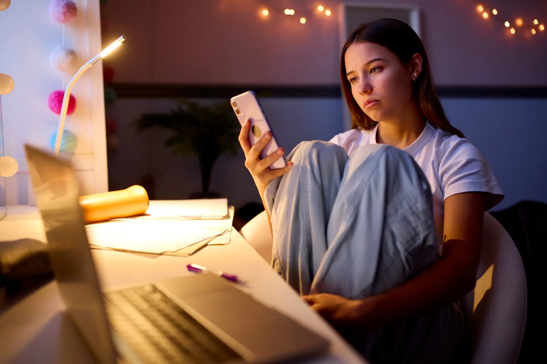 A young woman in pajamas sits at a desk with a laptop, lamp, and papers, looking thoughtfully at her phone. Warm fairy lights decorate the background, creating a cozy evening atmosphere.