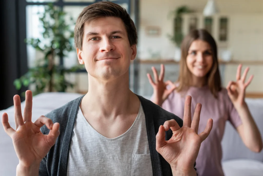 Un hombre y una mujer sonríen a la cámara, ambos haciendo el gesto de "OK" con la mano. Están en un interior y el fondo está enfocado suavemente, mostrando algunas plantas verdes y la decoración del hogar.