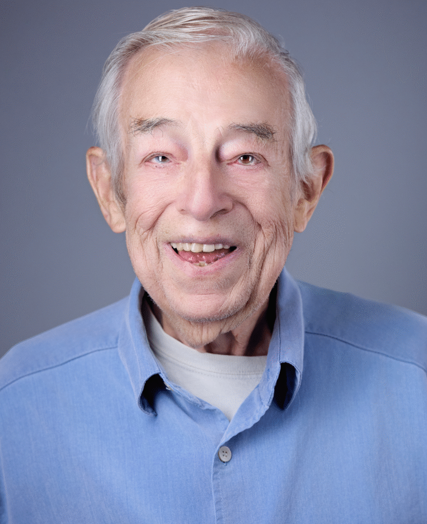 An elderly man with gray hair smiles warmly at the camera. He wears a light blue button-up shirt over a white undershirt, with a plain gray background—reflecting hope promoted by nonprofit supporting mental health.
