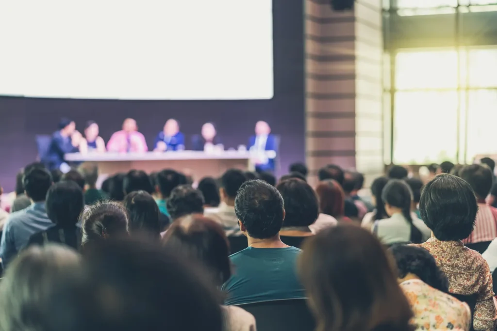 A large audience sits facing a stage where five people are seated at a panel table, engaging in a discussion in a well-lit conference room. The focus is on the audience from behind.